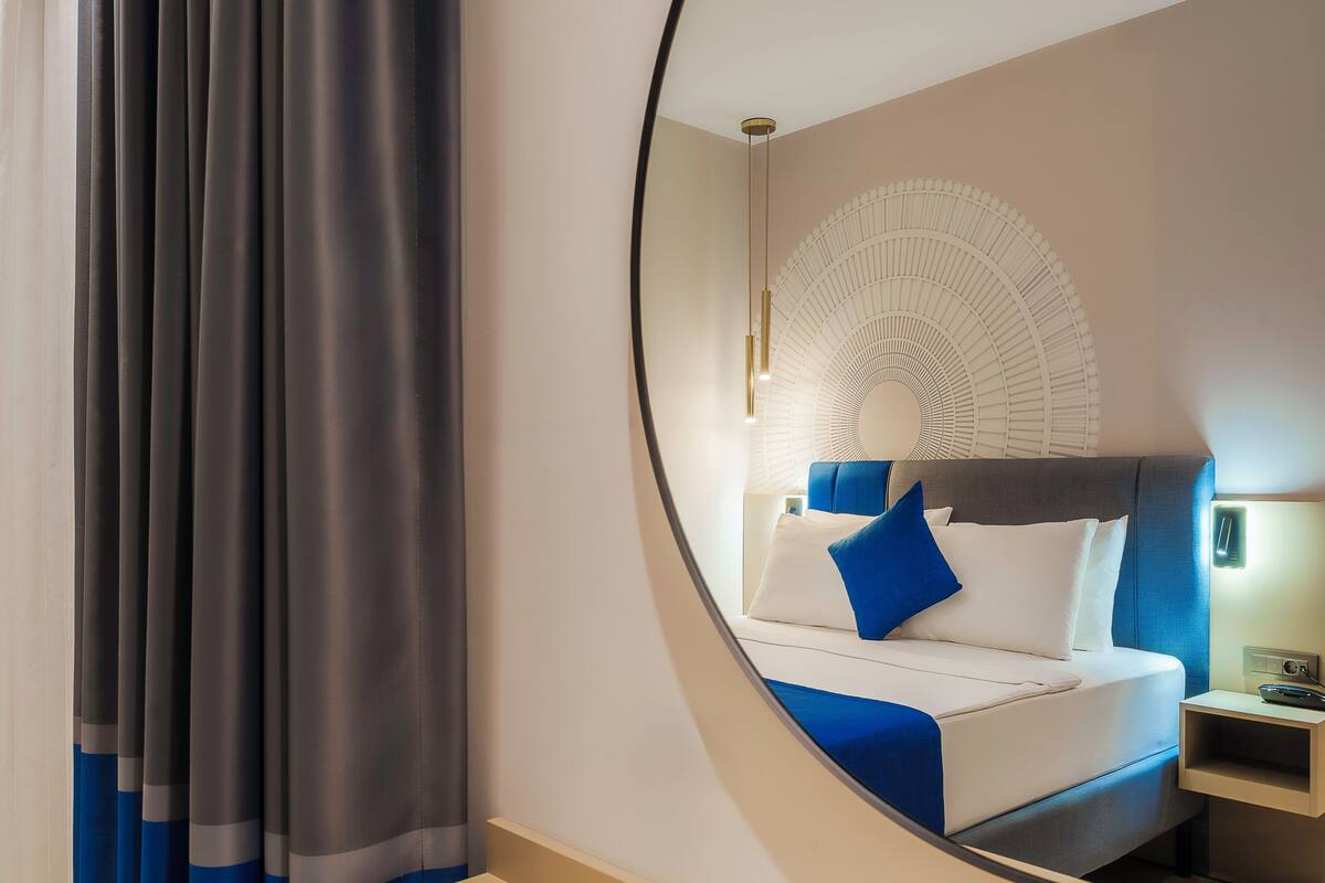 A modern hotel room with a bed, a nightstand, a mirror, and a large window with curtains.