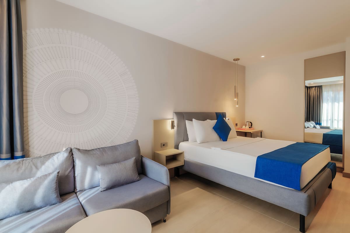 A modern hotel room with a bed, a sofa, a bedside table, a lamp, and a large abstract wall decoration.