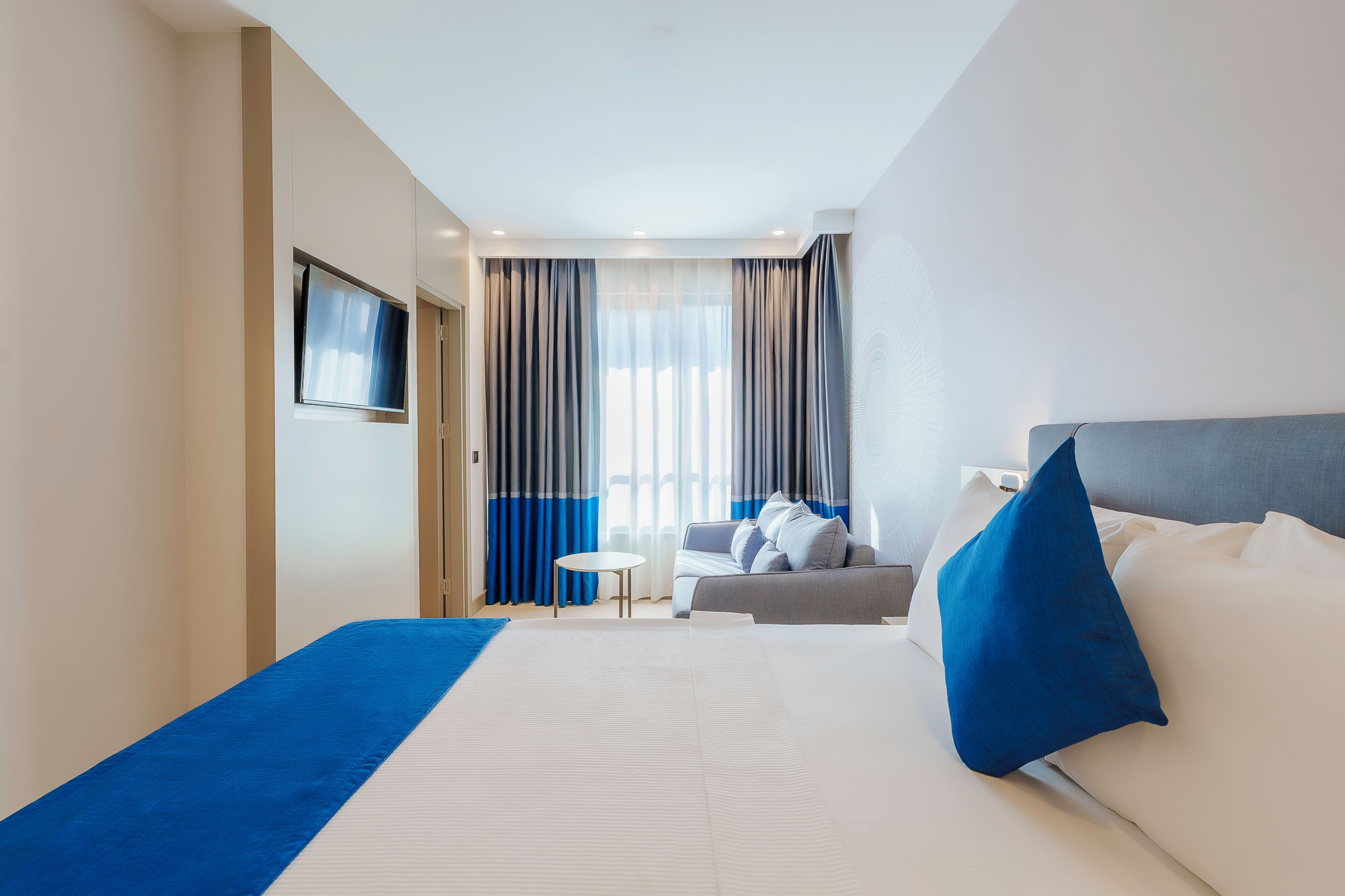 Quarto de hotel com cama, sofá, televisão e janela com cortinas.