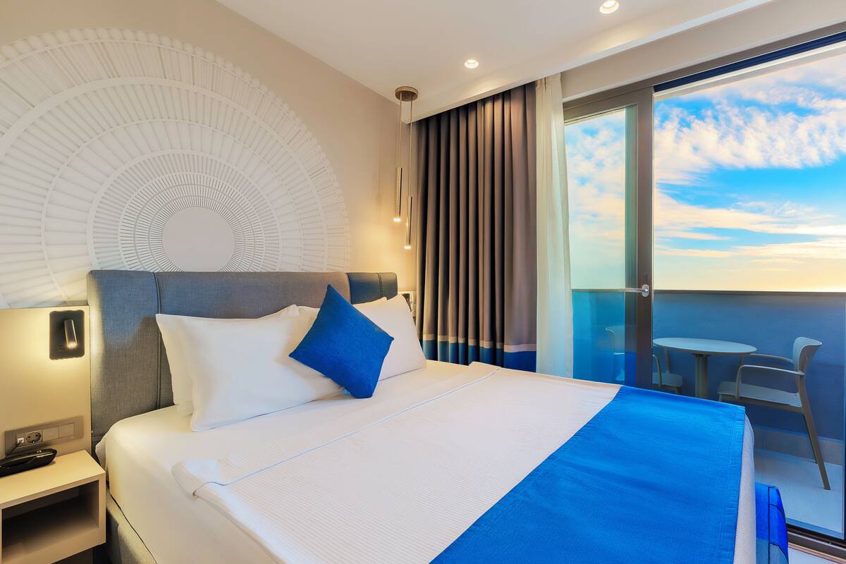 A modern hotel room with a large bed, a nightstand, a desk, and a balcony with a view of the sky.