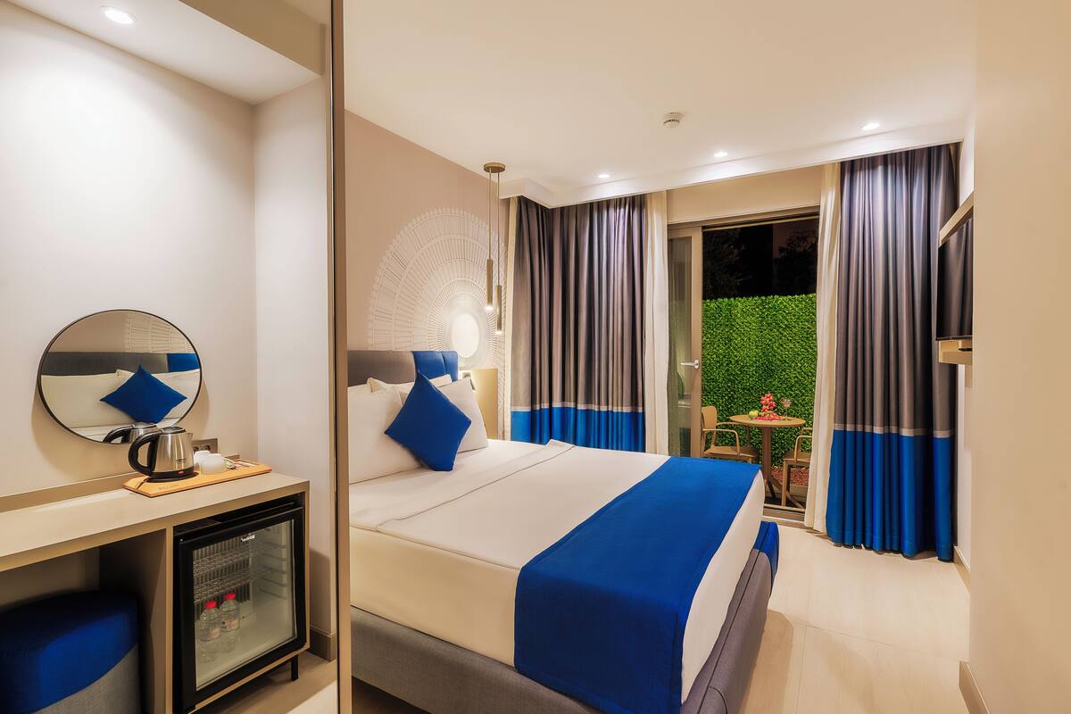 A hotel room with a bed, a desk with a coffee maker, a mini-fridge, and a view of a garden through the window.