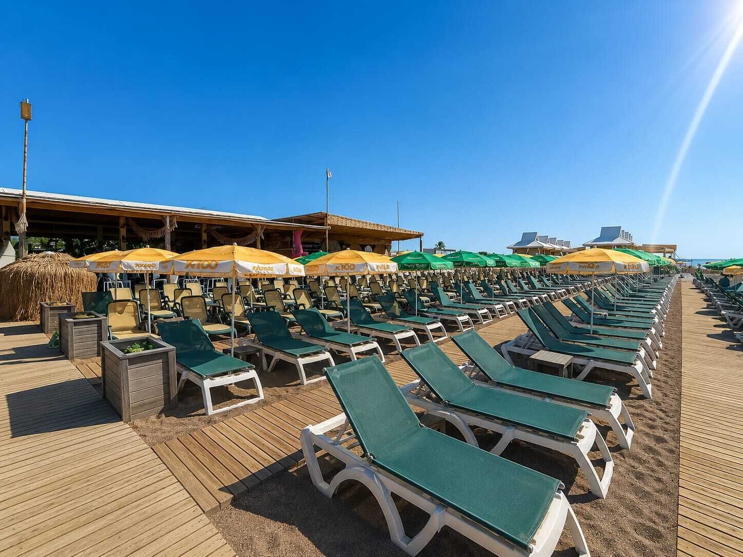 Un espace de détente aménagé au bord de la plage, avec des rangées de transats et de parasols, une terrasse en bois et un ciel dégagé.