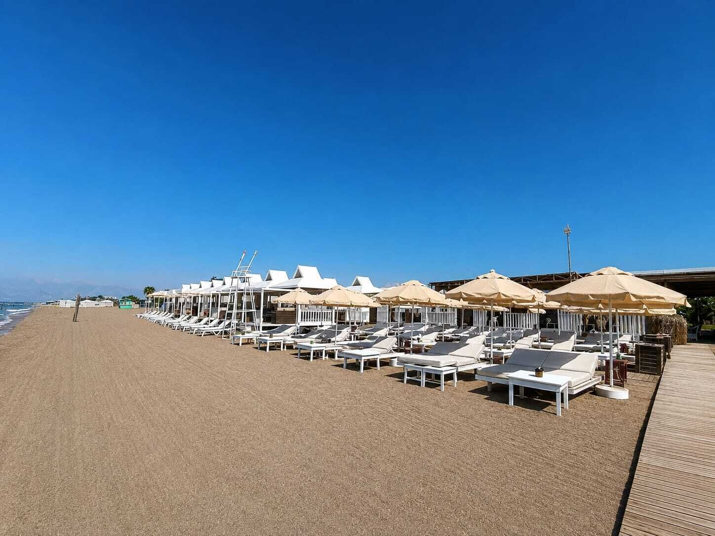 A beach with rows of sun loungers and umbrellas, a wooden walkway, and a clear blue sky.