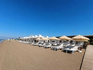 Une plage bordée de rangées de chaises longues et de parasols, une promenade en bois et un ciel d’un bleu limpide.