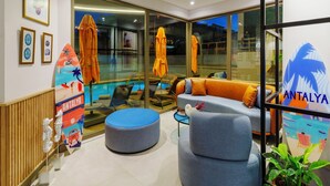 Een moderne hotellobby met een zwembadgedeelte, een zithoek met een bank en een poef, en ingelijste kunstwerken aan de muren.