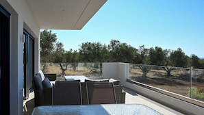 Un balcon doté d’une table en verre et de chaises en osier, donnant sur un espace aménagé avec des arbres et une clôture.