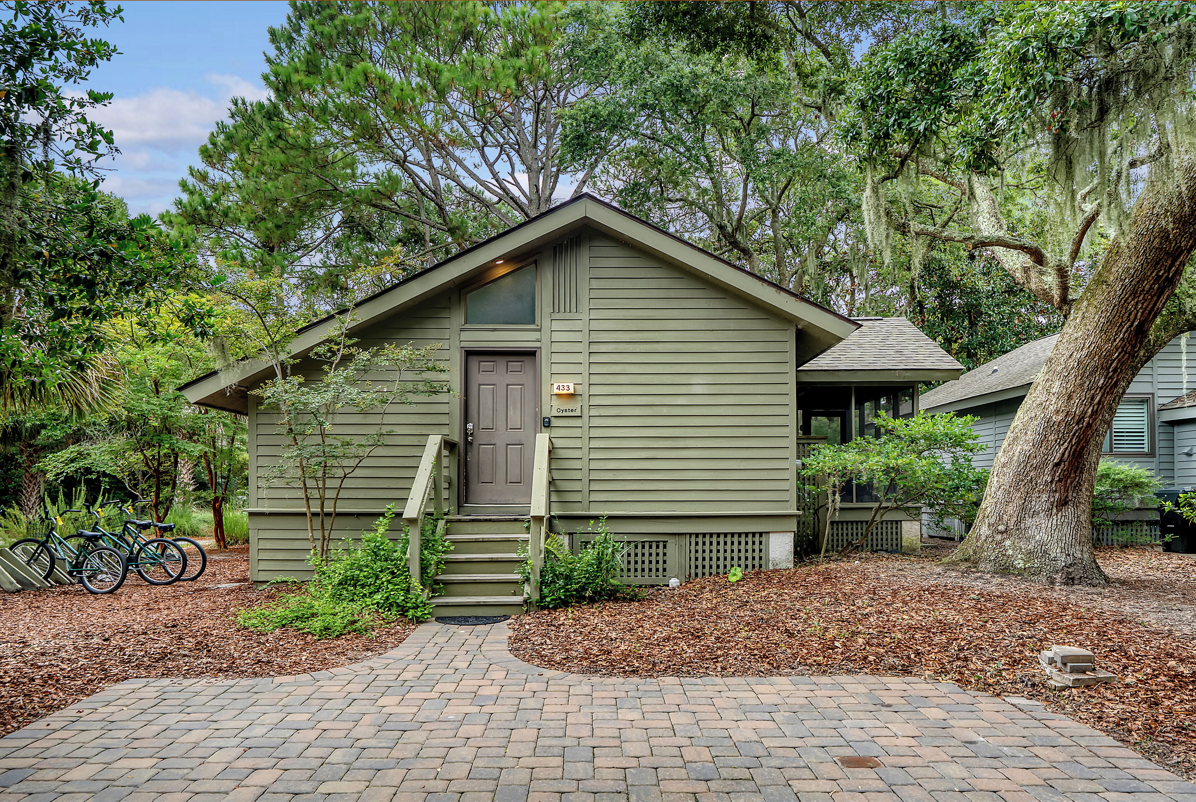 A small, single-story cabin with a front door and steps leading up to it, surrounded by trees and a brick pathway.