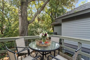 A round glass table with a wicker basket containing fruits and a vase of flowers, surrounded by chairs on a wooden deck. - Yards to the beach, charming cottage (Kiawah Island)