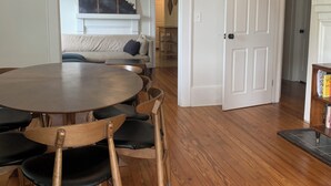 A dining area with a round table and wooden chairs, a ceiling fan, and a hallway leading to a living room with a sofa and a fireplace. - 2:1 Bunkroom Bungalow (Greenville)