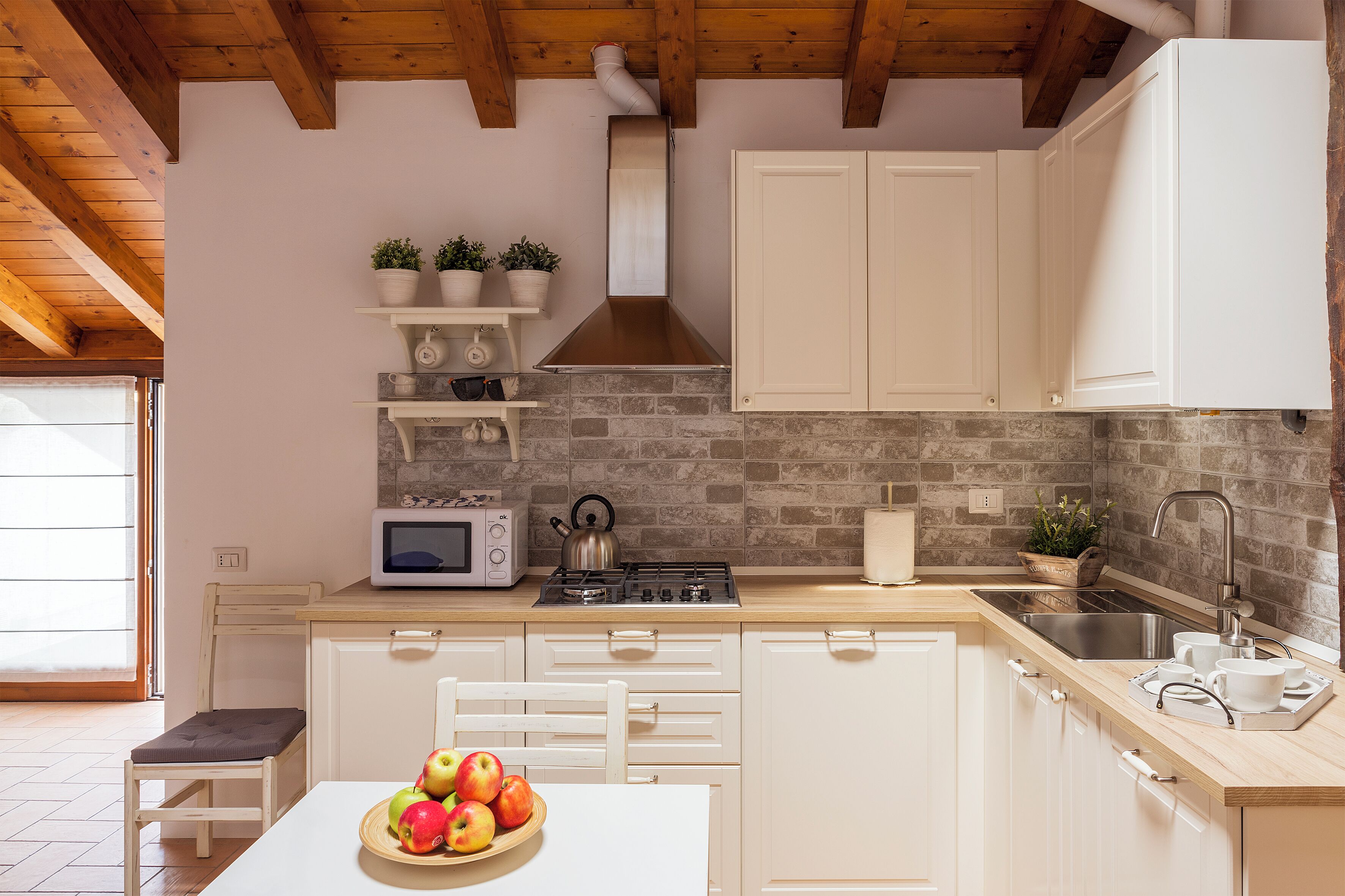 A kitchen with white cabinets, a microwave, a stove, and a sink. There are potted plants on a shelf and a bowl of fruit on the table.