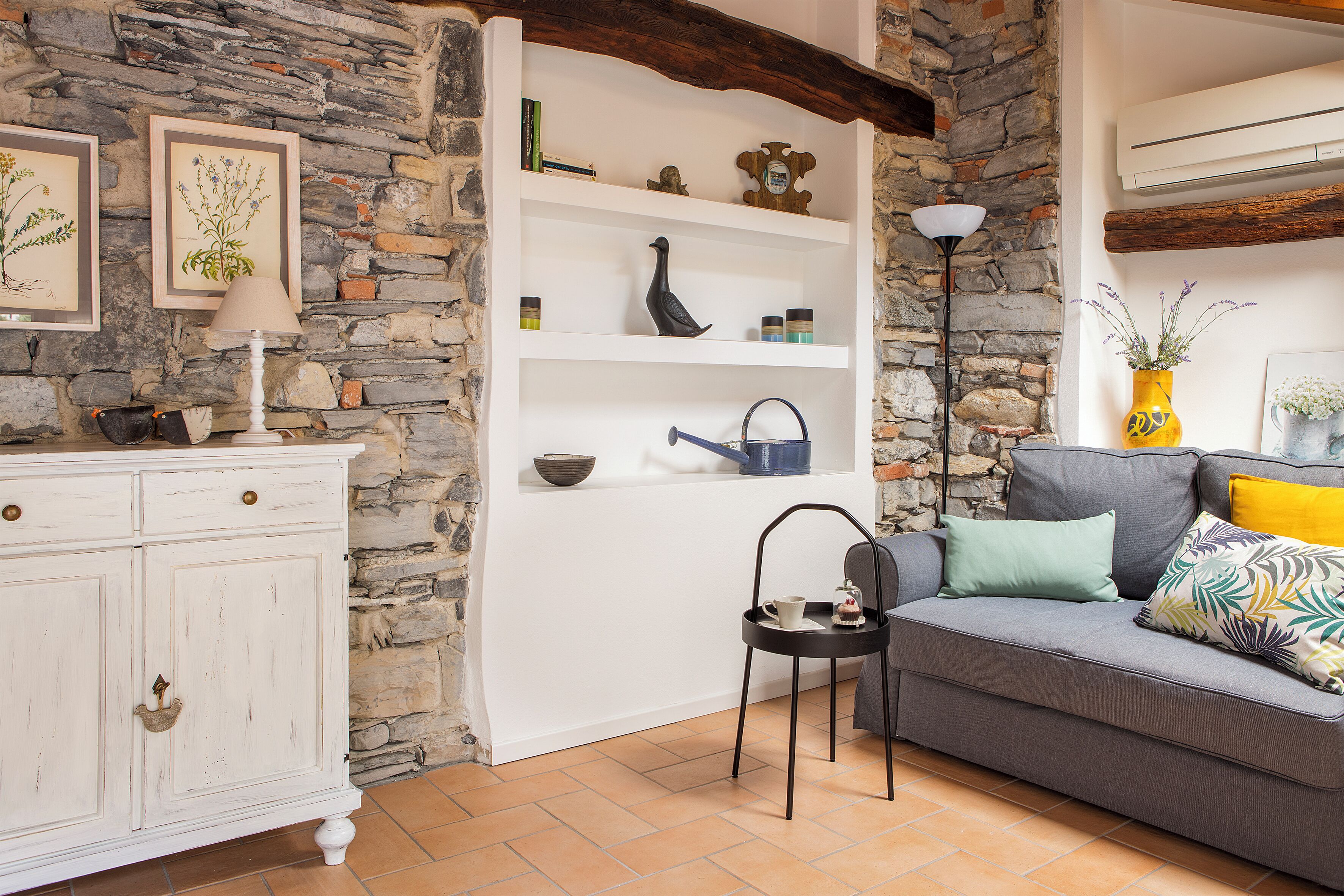 A cozy living room with a stone wall, a white cabinet, a grey sofa with cushions, and a small side table.