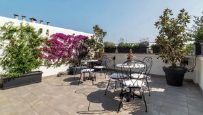 Eine Terrasse im Freien mit Tischen und Stühlen, umgeben von Blumentöpfen und einer weißen Mauer.