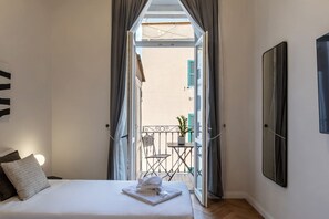 A bedroom with a bed, a nightstand, a lamp, a window with curtains, a balcony with a chair and a plant, and a wall-mounted mirror.