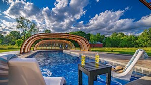 Una piscina con sillas de descanso y una mesa, rodeada por una zona de estar cubierta y vistas a árboles y un cielo despejado.