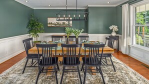 Comedor con mesa de madera, sillas negras, una lámpara de araña y vistas a zonas verdes al exterior.
