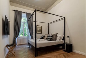A four-poster bed with a white mattress and patterned pillows, a black nightstand with a lamp, and a window with curtains. - Castro Pretorio 9 - FromHometoRome (Roma)