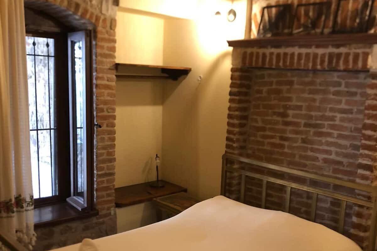 A bedroom with a bed, a brick fireplace, a window with curtains, and a wooden ceiling.