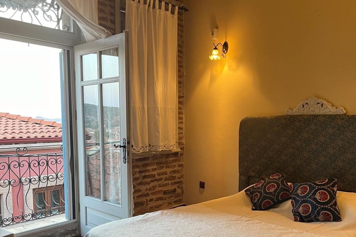 A bedroom with a bed, a nightstand, a window with curtains, a balcony view, and a wall-mounted light fixture.