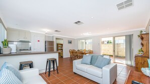 A spacious living area with a white sofa, a dining table with chairs, and a kitchen area with cabinets and a refrigerator. - Beauty And the Beach - Coastal Comfort at its best. (Guilderton)