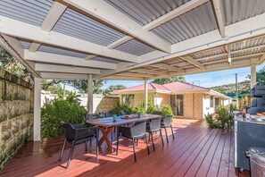 An outdoor patio with a wooden table and chairs, a grill, and a covered area with a view of a red-roofed building in the background. - Beauty And the Beach - Coastal Comfort at its best. (Guilderton)