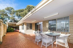 A modern outdoor patio with a glass table and wicker chairs, surrounded by stone walls and large windows. - Beauty And the Beach - Coastal Comfort at its best. (Guilderton)