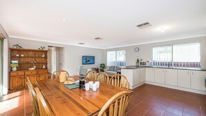 A spacious kitchen with white cabinets, a wooden dining table, and a large window providing natural light. - Beauty And the Beach - Coastal Comfort at its best. (Guilderton)