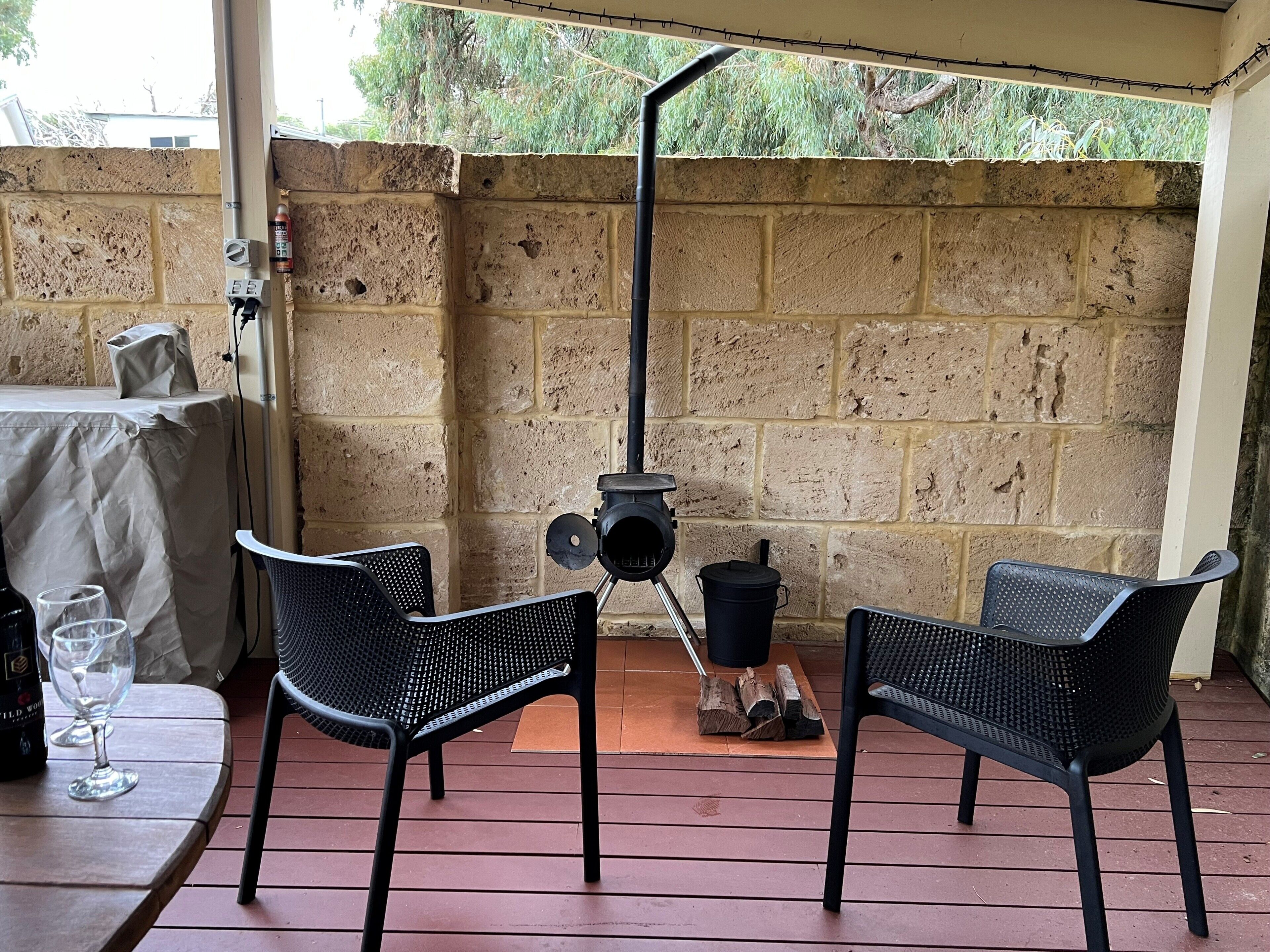 Un patio al aire libre con dos sillas de malla negra, una mesa con una botella y un vaso, espacio para leña y un muro de ladrillo.