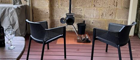 Un patio al aire libre con dos sillas de malla negra, una mesa con una botella y un vaso, espacio para leña y un muro de ladrillo.