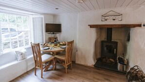 A cozy dining area with a wooden table and chairs, a television, and a wood-burning stove. - Tamarisk Cottage (St. Agnes)