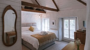A bedroom with a bed, a wooden headboard, a mirror, a bedside table, a dresser, a window with blinds, and a ceiling light. - Tamarisk Cottage (St. Agnes)