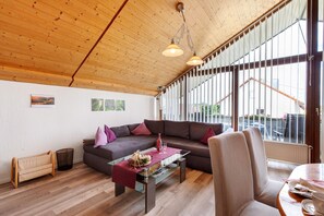 Una sala moderna con techo de madera, una mesa de centro de vidrio, un sofá marrón y ventanas amplias.