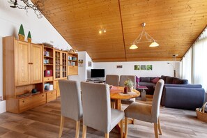 Un salón amplio con techo de madera, una mesa de comedor con sillas, un sofá y un televisor.