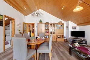Un amplio salón con techo de madera, una mesa de comedor con sillas, una estantería, un televisor y una cocina visible a través de una puerta abierta.