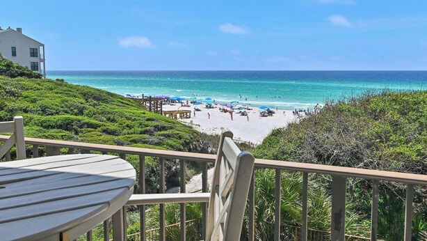 Blick auf einen Strand vom Balkon aus, mit Tisch und Stühlen, auf einen Sandstrand mit Sonnenschirmen und Menschen.