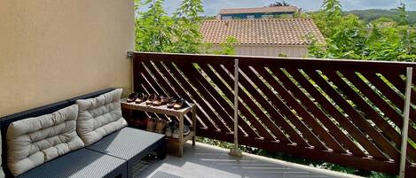 Un área de estar al aire libre con techo, que incluye un sofá, una mesa de centro, una valla de madera y vistas a edificios y árboles.