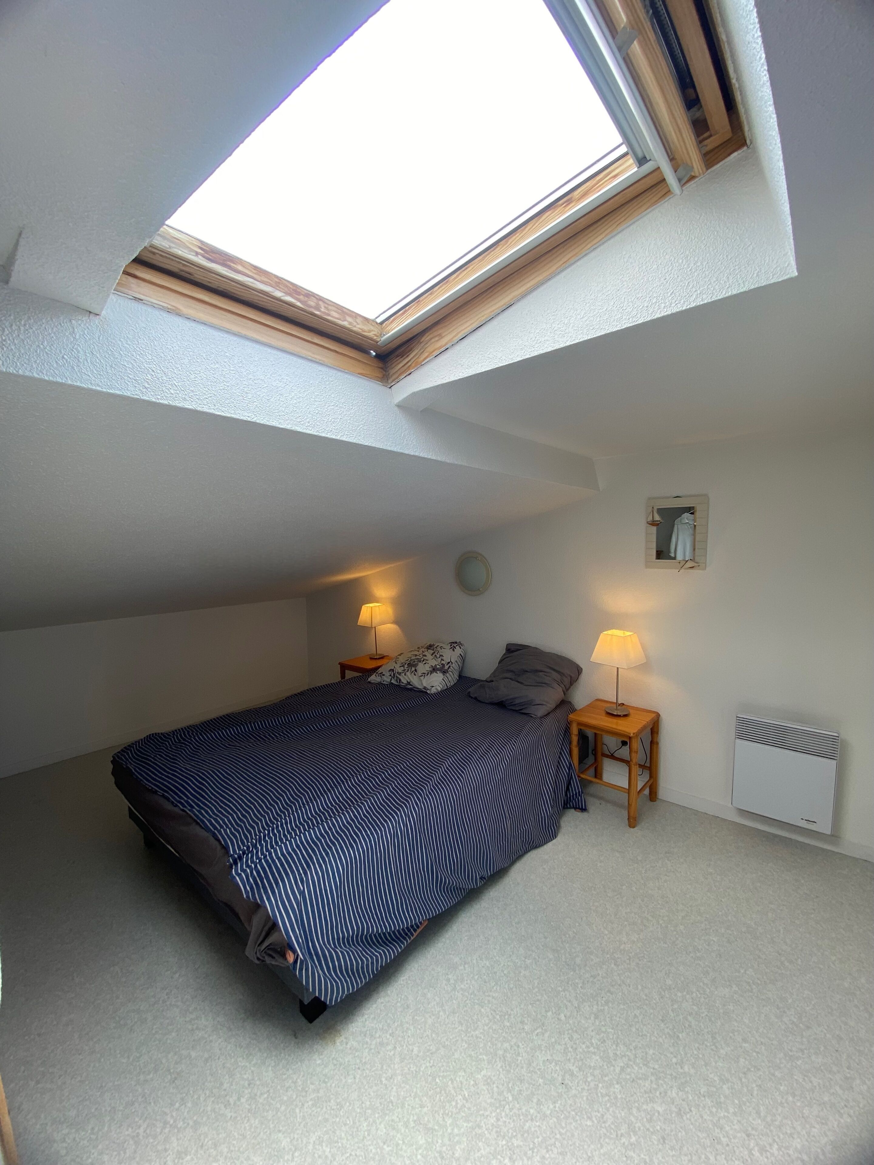 Ein Schlafzimmer mit Dachfenster, einem Bett, zwei Nachttischen mit Lampen, einem Heizkörper und einem kleinen Wandspiegel.