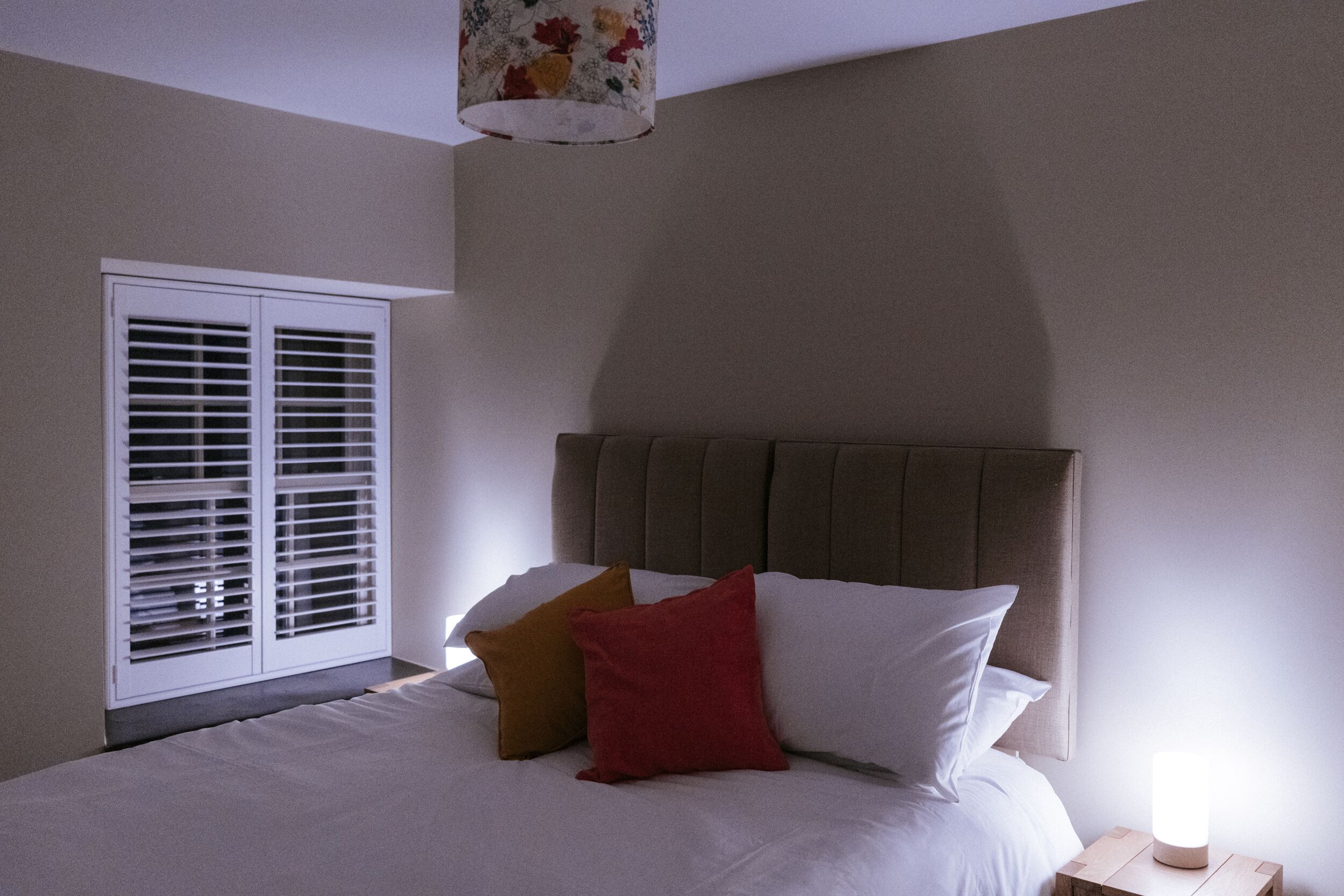 A bedroom with a bed, pillows, a nightstand, a lamp, and a window with blinds.