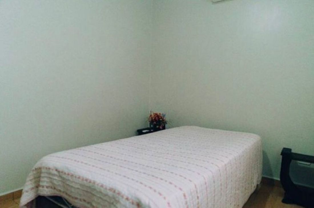 Una habitación pequeña y poco amoblada, con una cama individual, una mesita con un florero y una silla.