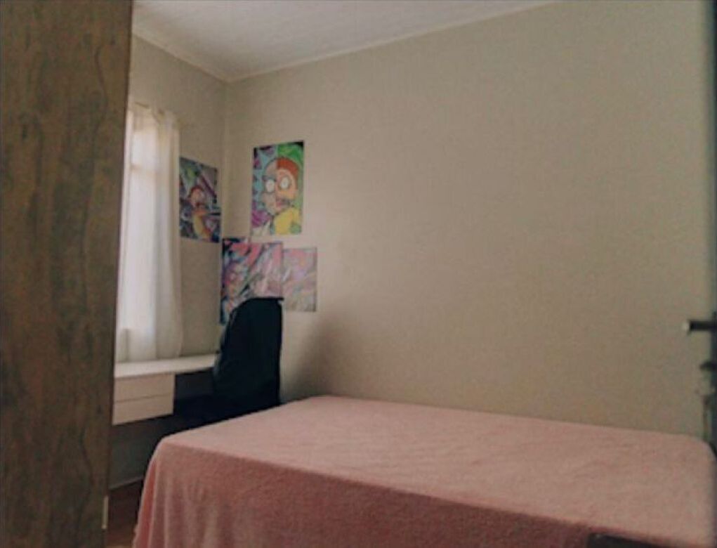 A room with a bed, a desk, and a chair. There are two colorful paintings on the wall.