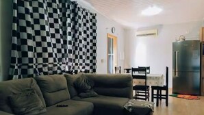 Olohuone, jossa on sohva, ruokapöytä tuoleineen, jääkaappi ja ruutukuviollinen verho.