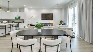 Eine moderne Küche mit weißen Schränken, ein Essbereich mit einem runden Tisch und Stühlen, eine Sofagarnitur und ein großes Fenster mit Vorhängen.