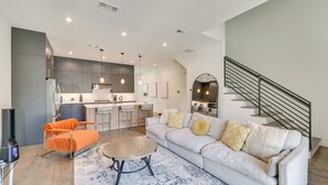A modern living room with a sofa, coffee table, and orange armchair. A kitchen with bar stools is visible in the background. - Modern & Spacious Home 3 Mi to Downtown Dallas! (Dallas)
