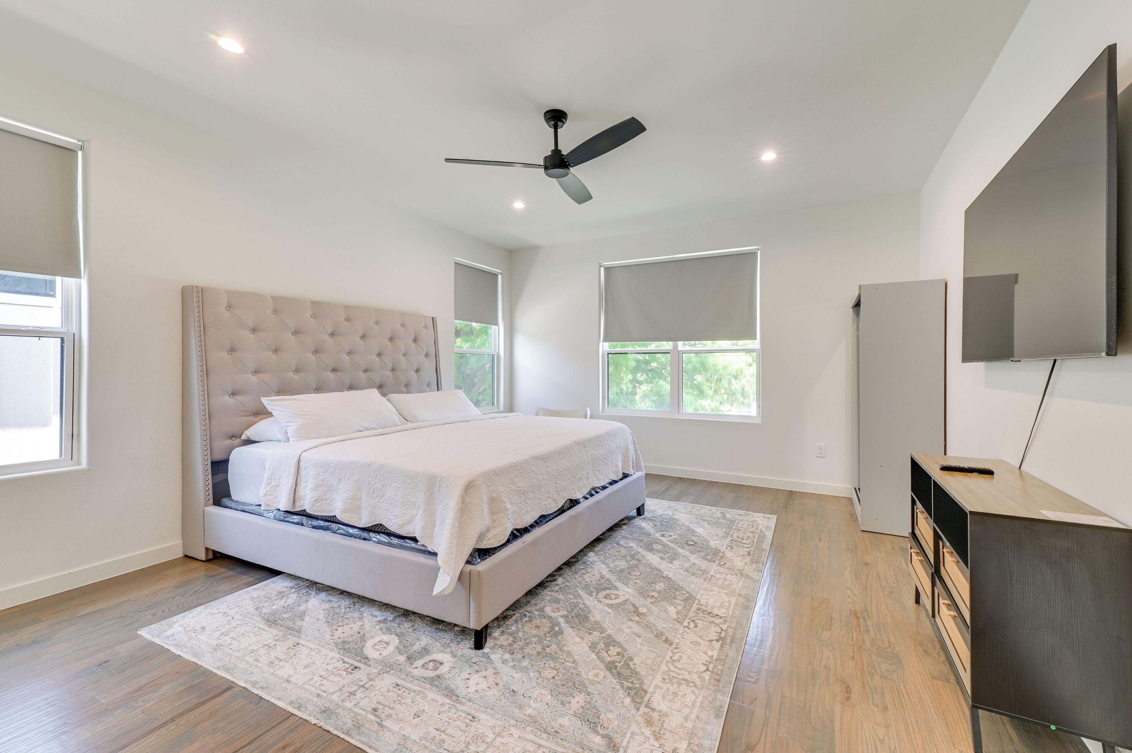 Un dormitorio con cama y cabecera capitonada, ventilador de techo, ventanas amplias, televisor de pantalla plana y cómoda de madera.