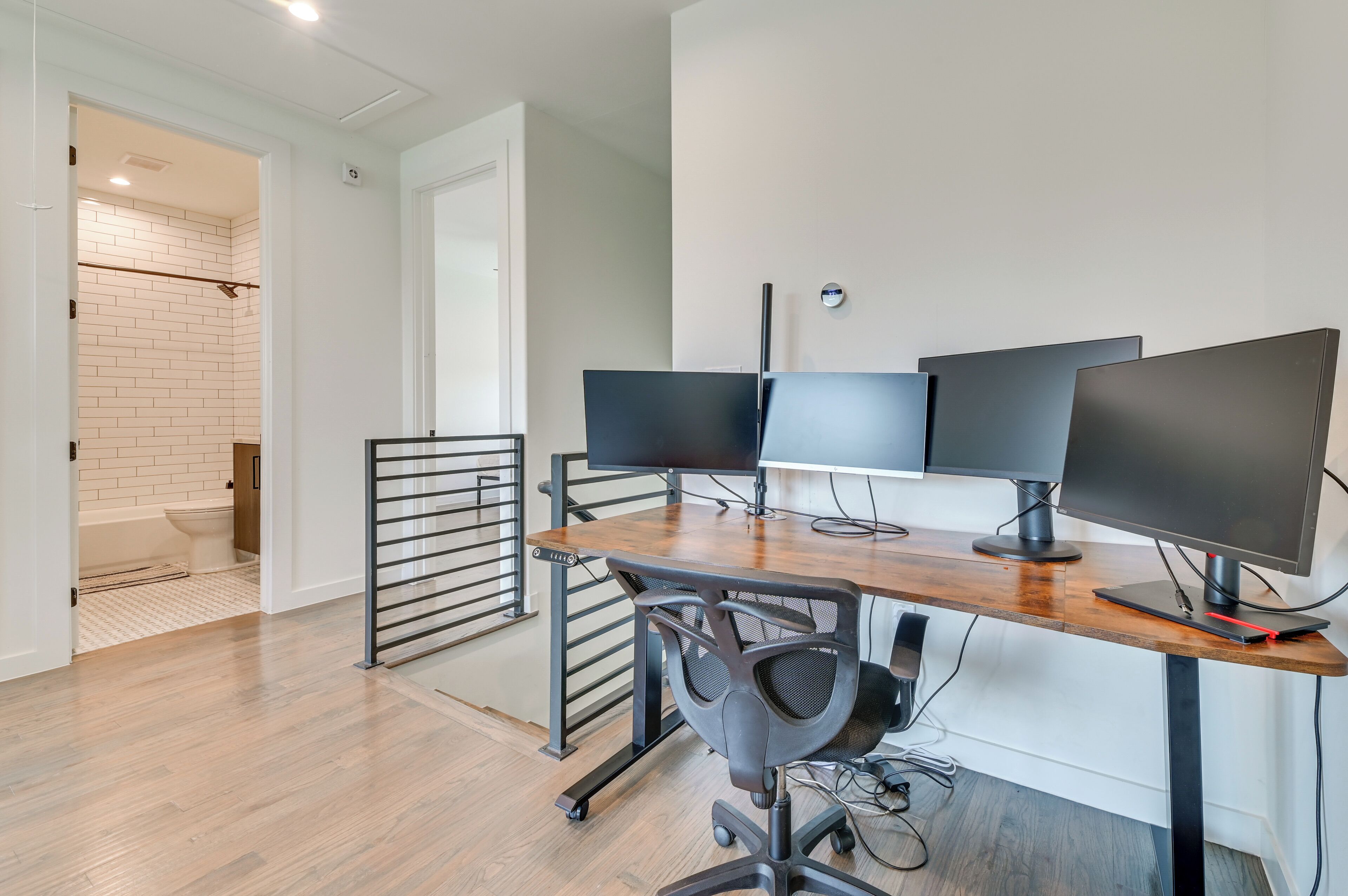 Un espace de bureau moderne avec un bureau en bois, trois écrans, un fauteuil de bureau noir et un mur en briques blanches.