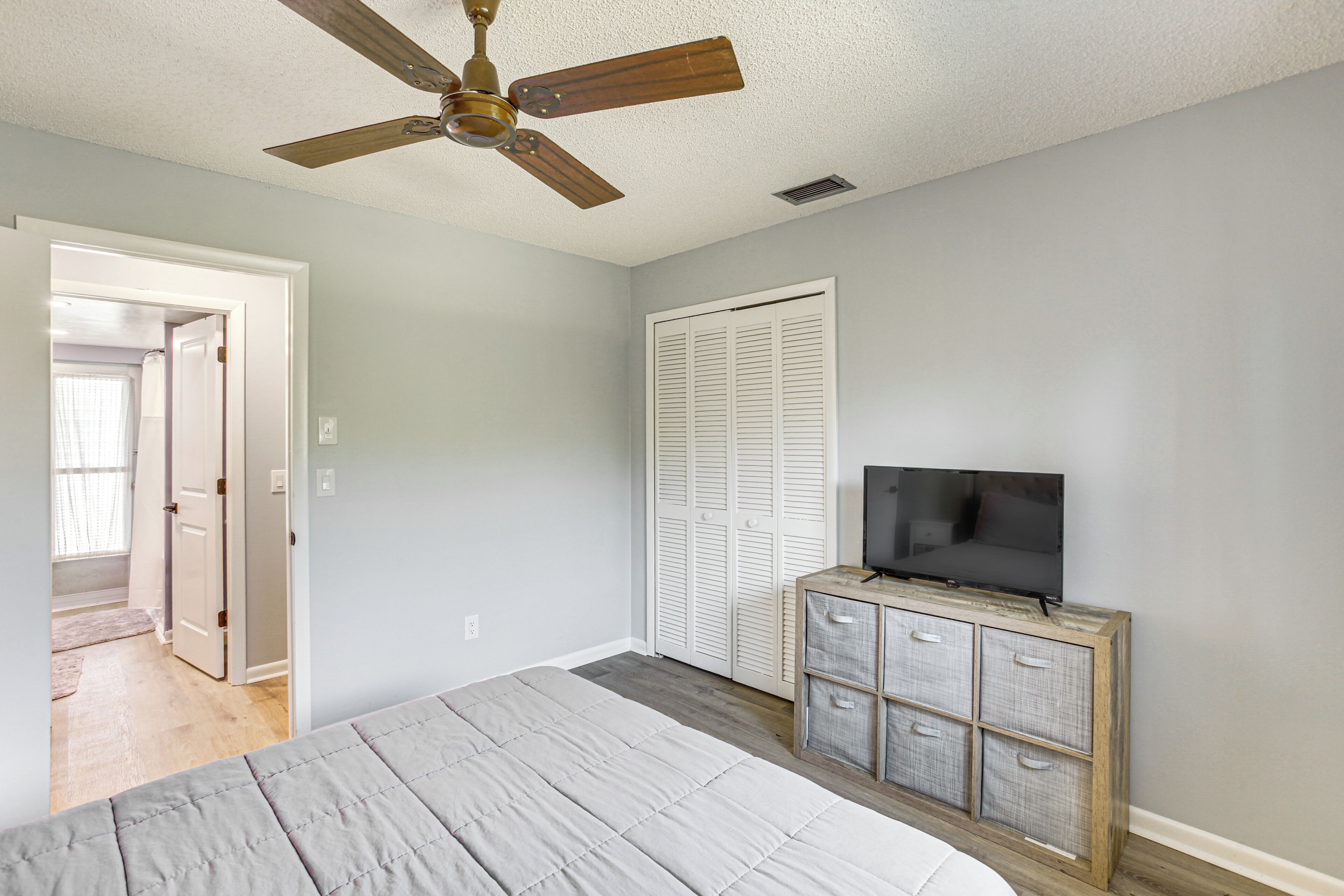 Une chambre à coucher avec un lit, un ventilateur de plafond, un téléviseur posé sur une commode en bois et une porte donnant sur une autre pièce.