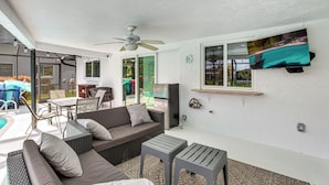 A covered outdoor living area with a sofa, two tables, a ceiling fan, and a TV mounted on the wall. - Swim, Hike & Golf: Family Gem in Cape Coral! (Cape Coral)