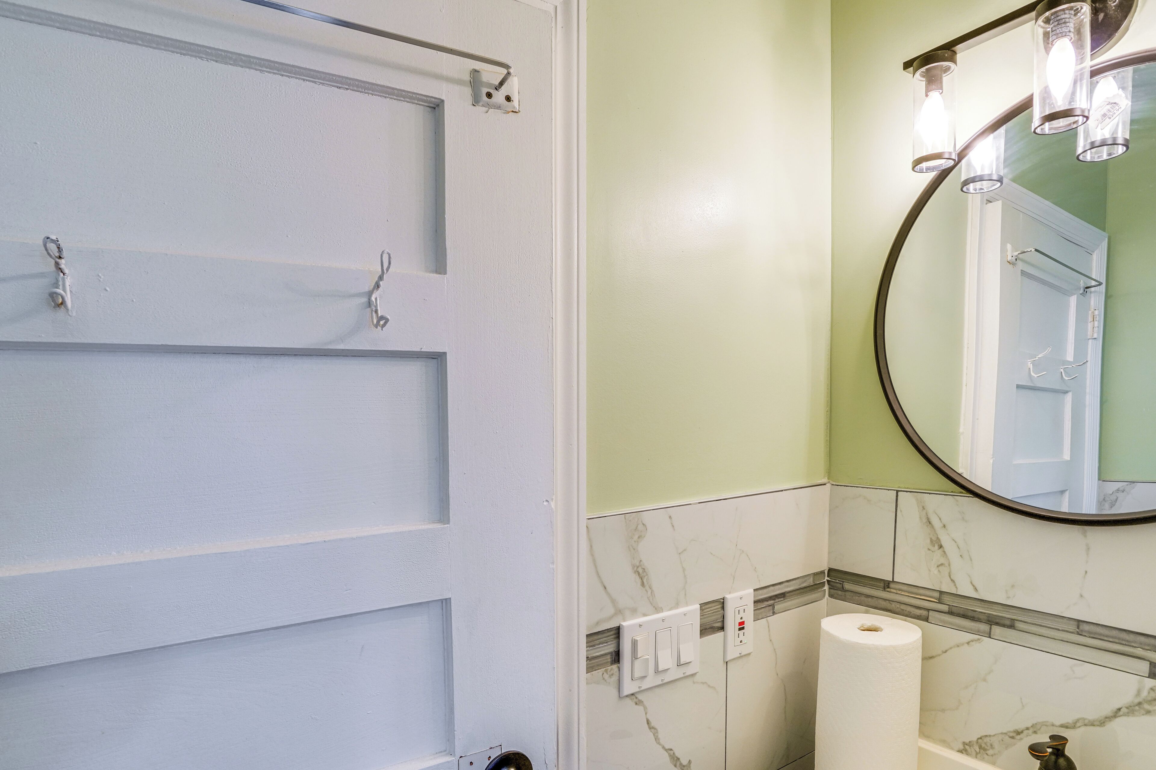 A bathroom with a white door, a marble-tiled wall, a round mirror with three lights, and a toilet paper roll on the floor.