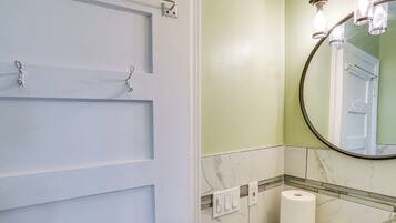 A bathroom with a white door, a marble-tiled wall, a round mirror with three lights, and a toilet paper roll on the floor.