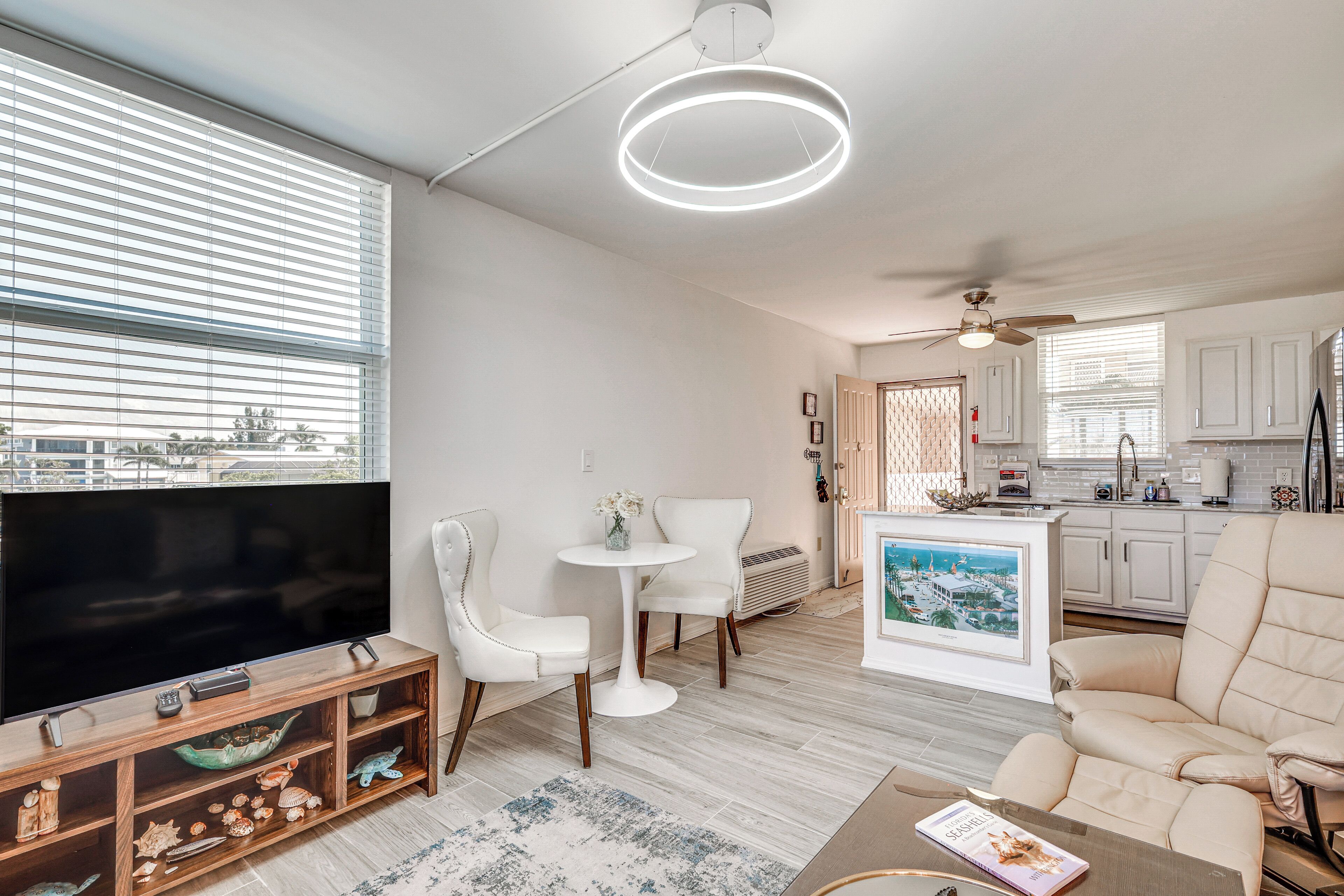 A modern living room with a flat-screen TV, white furniture, a small round table, and a kitchen area with white cabinets and a tiled backsplash.