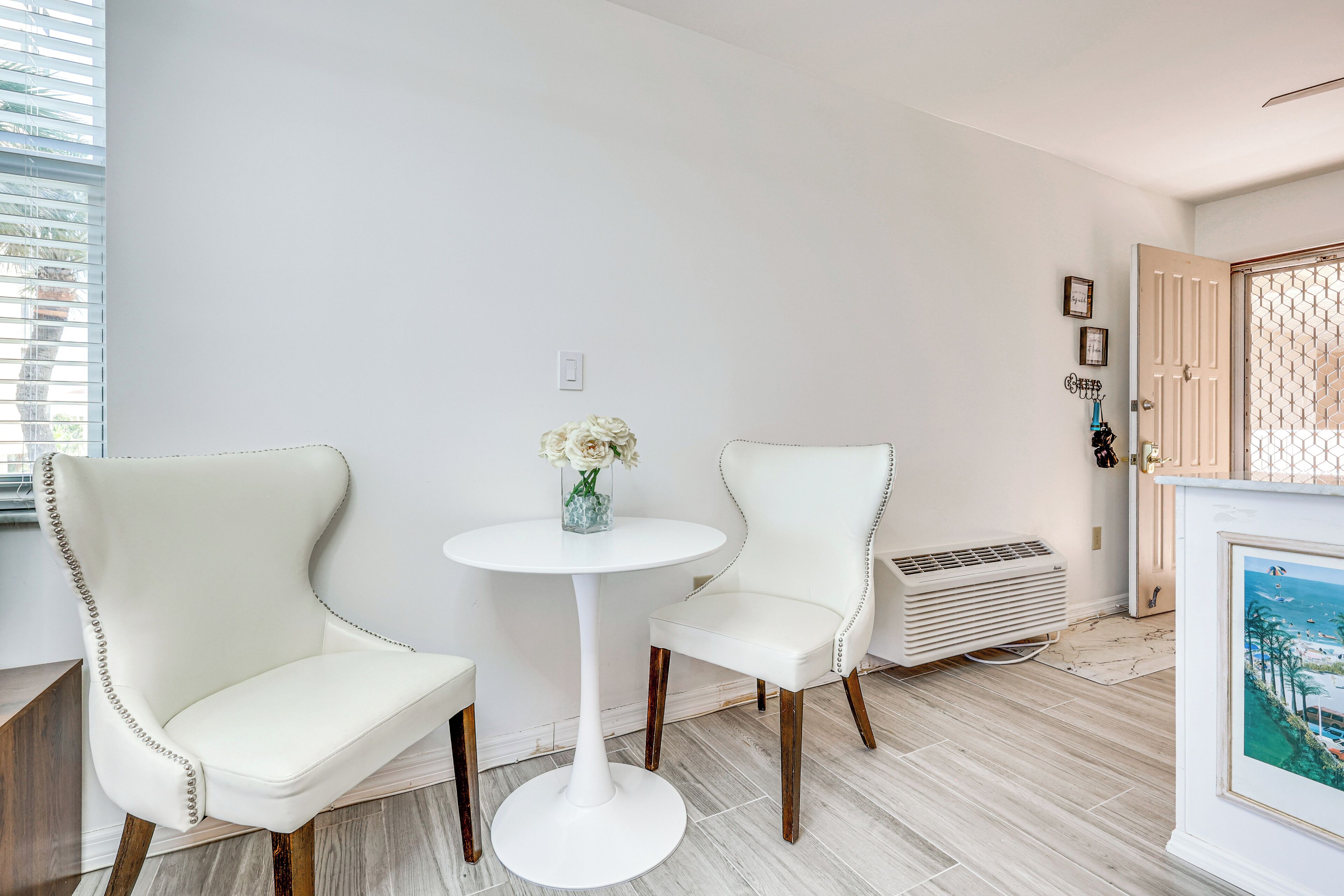 A room with two white upholstered chairs, a round white table, and a small vase with flowers.